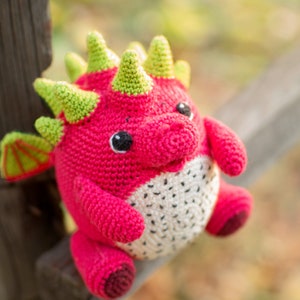 Op de afbeelding: Een gehaakt draken speelgoed met roze en groene schubben, witte en zwarte vlekken en zwarte ogen. De draak zit op een houten hek.