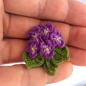 May include: A small crocheted bouquet of purple flowers with green leaves. The flowers have yellow centers.