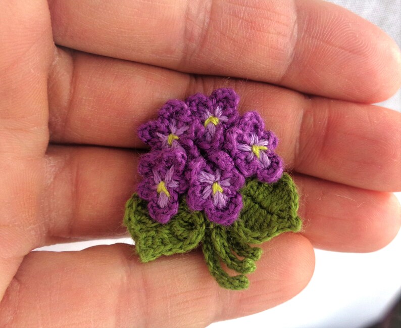 May include: A small crocheted bouquet of purple flowers with green leaves. The flowers have yellow centers.