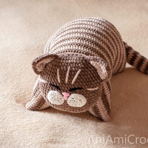 May include: A crocheted cat toy featuring brown and beige horizontal stripes across its body and tail. The cat has a brown head with white muzzle patches, a small pink nose, and closed eyes. The text AniAmiCrochet is visible in the bottom right corner.