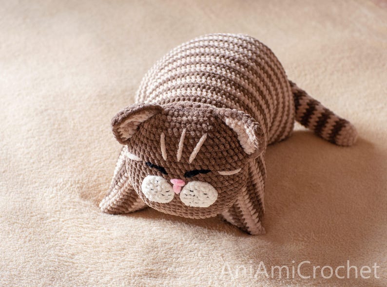May include: A crocheted cat toy featuring brown and beige horizontal stripes across its body and tail. The cat has a brown head with white muzzle patches, a small pink nose, and closed eyes. The text AniAmiCrochet is visible in the bottom right corner.