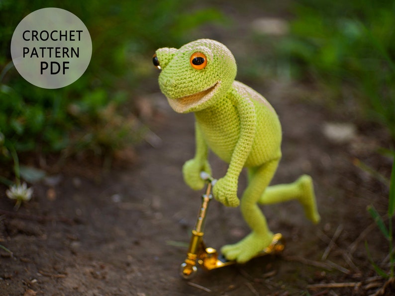 May include: A green crocheted frog wearing orange eyes is riding a gold scooter on a dirt path. The frog has a wide smile and is looking at the camera. The image includes the text "CROCHET PATTERN PDF".