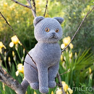 May include: A handmade gray crochet cat with orange eyes sits on a tree branch. The plush cat has a textured surface and is surrounded by yellow flowers and greenery. The cat is a whimsical, handcrafted item.