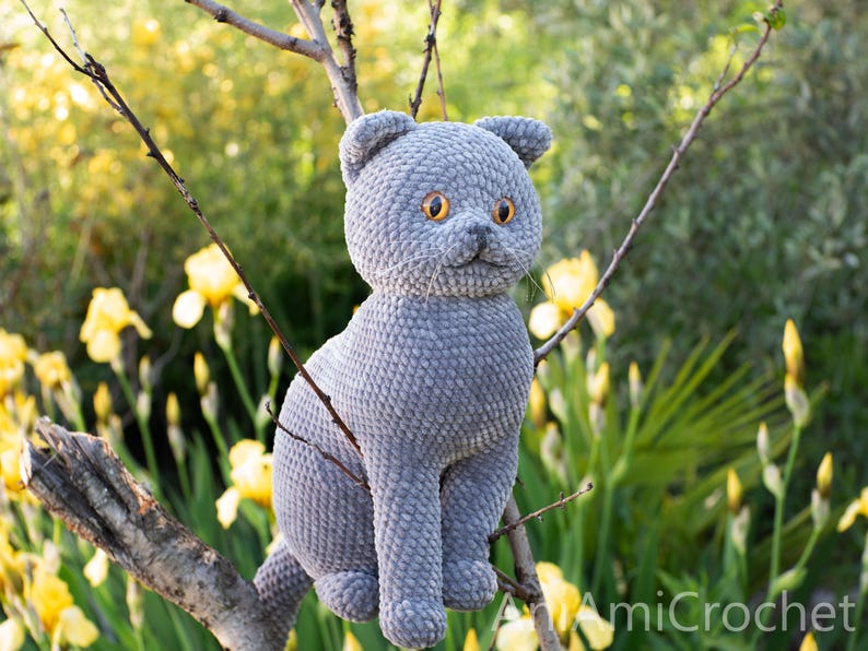 May include: A handmade gray crochet cat with orange eyes sits on a tree branch. The plush cat has a textured surface and is surrounded by yellow flowers and greenery. The cat is a whimsical, handcrafted item.