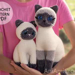 May include: Two Siamese cat plush toys made of soft yarn, featuring cream-coloured bodies, dark grey ears, faces, and paws, with bright blue eyes. The image includes the text CROCHET PATTERN PDF and AniAmiCrochet. The cats are held in front of a pink shirt.