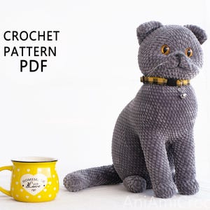 May include: A plush gray cat with a yellow and black collar and a small bell. The cat is sitting upright. A yellow mug with white polka dots and the text "Homemade with Love" is to the left. The text "CROCHET PATTERN PDF" is at the top.