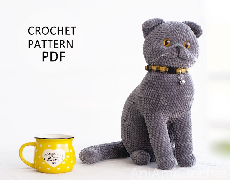 May include: A plush gray cat with a yellow and black collar and a small bell. The cat is sitting upright. A yellow mug with white polka dots and the text "Homemade with Love" is to the left. The text "CROCHET PATTERN PDF" is at the top.