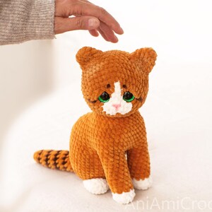 May include: A plush crochet cat toy made of soft orange yarn with a white chest, white paws, and a striped tail. The cat features large, bright green plastic eyes, a small pink stitched nose, and pointed ears, sitting in an upright position.