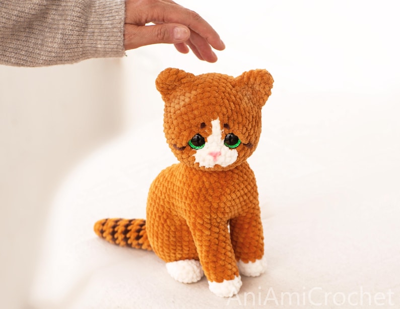 May include: A plush crochet cat toy made of soft orange yarn with a white chest, white paws, and a striped tail. The cat features large, bright green plastic eyes, a small pink stitched nose, and pointed ears, sitting in an upright position.