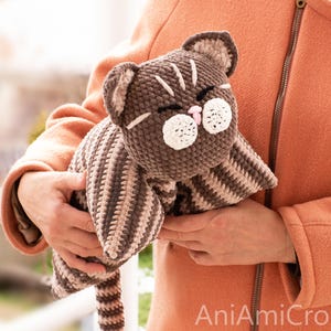 May include: A brown and beige striped crochet cat plush toy with a round head, pointed ears, closed eyes, and white circular muzzle patches. The cat has a small pink nose and a long striped tail. The text AniAmiCrochet is visible in the corner.
