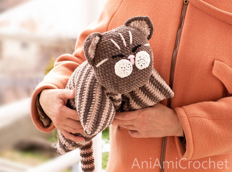 May include: A brown and beige striped crochet cat plush toy with a round head, pointed ears, closed eyes, and white circular muzzle patches. The cat has a small pink nose and a long striped tail. The text AniAmiCrochet is visible in the corner.