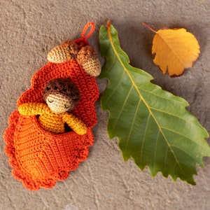 May include: A crocheted acorn with a sleeping doll inside. The acorn is orange and brown and the doll is wearing a yellow and orange outfit. The acorn is sitting on a gray surface with a green leaf.