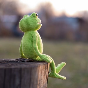 May include: A green crocheted frog sitting on a tree stump. The frog is looking up and has a big smile.