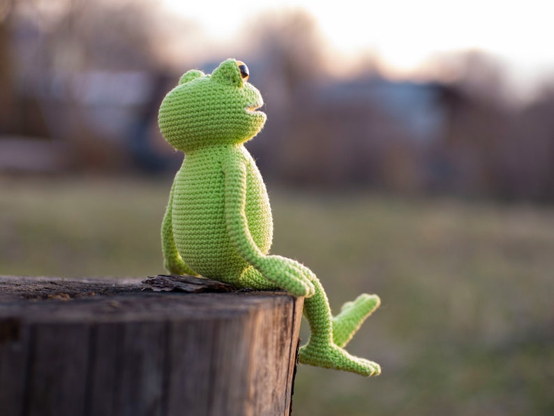 May include: A green crocheted frog sitting on a tree stump. The frog is looking up and has a big smile.