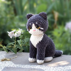 May include: A handmade crochet cat figurine featuring dark gray yarn for the body and head, with white yarn accents on the chest, paws, and face. The cat has large, glossy brown eyes, a small pink stitched nose, and pointed ears, crafted in a seated pose.