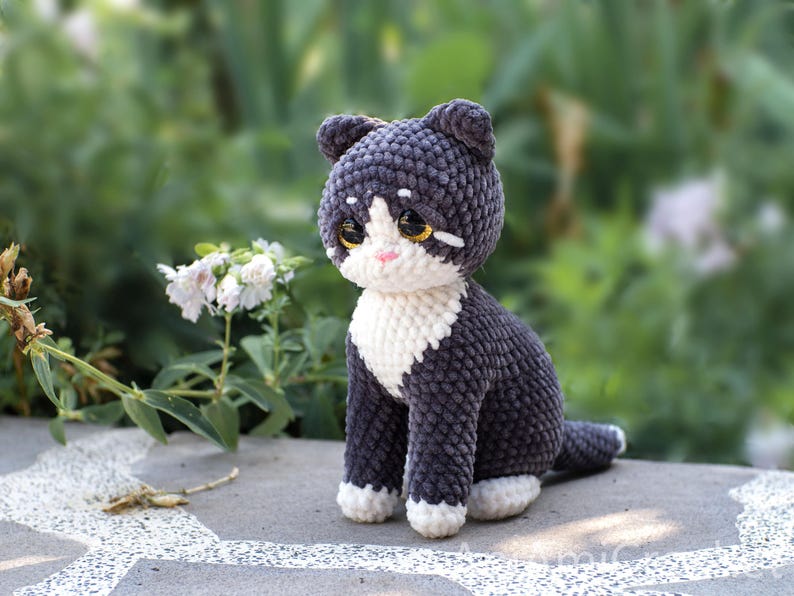 May include: A handmade crochet cat figurine featuring dark gray yarn for the body and head, with white yarn accents on the chest, paws, and face. The cat has large, glossy brown eyes, a small pink stitched nose, and pointed ears, crafted in a seated pose.