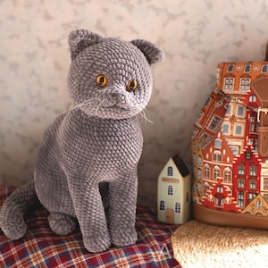 May include: A gray crocheted cat with yellow eyes sits on a red and brown plaid surface. A colorful backpack with a cityscape design and a small decorative house are in the background. The cat has a textured, plush appearance.