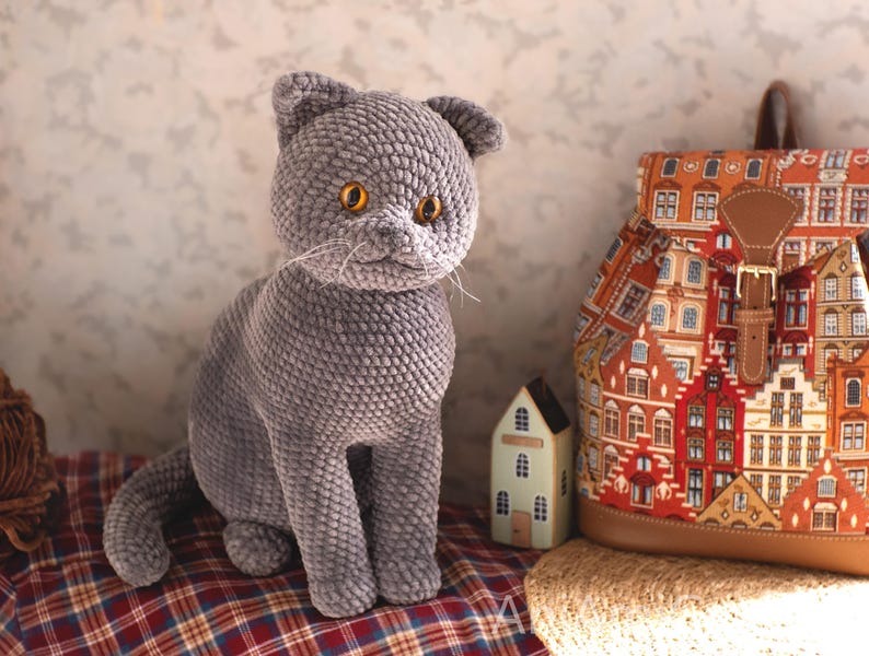 May include: A gray crocheted cat with yellow eyes sits on a red and brown plaid surface. A colorful backpack with a cityscape design and a small decorative house are in the background. The cat has a textured, plush appearance.