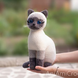 May include: A crocheted Siamese cat plush toy with a cream-colored body, dark gray ears, paws, and face mask. The cat features bright blue plastic eyes, stitched whiskers, and the text AniAmiCrochet printed in white in the bottom right corner.