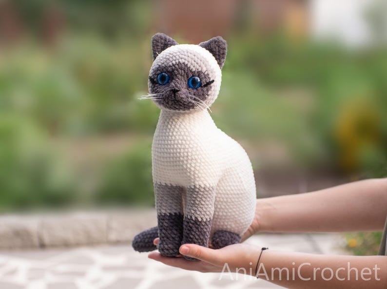 May include: A crocheted Siamese cat plush toy with a cream-colored body, dark gray ears, paws, and face mask. The cat features bright blue plastic eyes, stitched whiskers, and the text AniAmiCrochet printed in white in the bottom right corner.