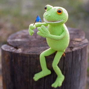 May include: A green crocheted frog toy sitting on a tree stump, holding a blue flower. The frog has a wide smile and orange eyes.