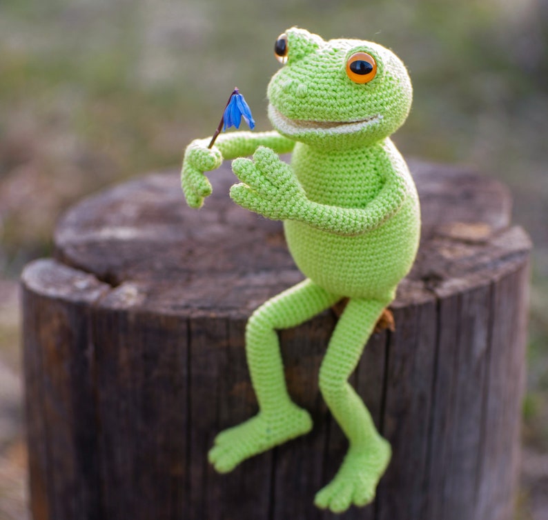 May include: A green crocheted frog toy sitting on a tree stump, holding a blue flower. The frog has a wide smile and orange eyes.