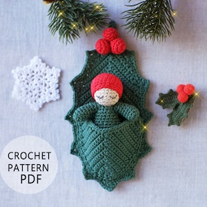 Può includere: Schema a uncinetto PDF per una foglia di agrifoglio verde con una bambola addormentata all'interno con un cappello rosso. La bambola ha gli occhi chiusi e un viso bianco. La foglia di agrifoglio ha bacche rosse nella parte superiore.