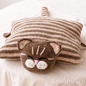 May include: A rectangular crochet cushion featuring a brown and beige striped pattern with a cat head attached to the front. The cat head is dark brown with white muzzle patches, a small pink nose, and embroidered eyes. The text AniAmiCrochet is visible.