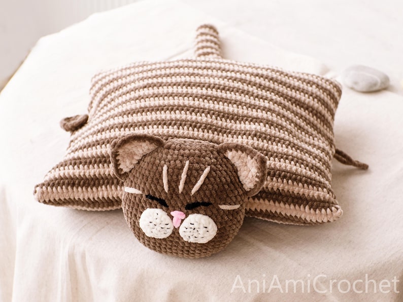 May include: A rectangular crochet cushion featuring a brown and beige striped pattern with a cat head attached to the front. The cat head is dark brown with white muzzle patches, a small pink nose, and embroidered eyes. The text AniAmiCrochet is visible.