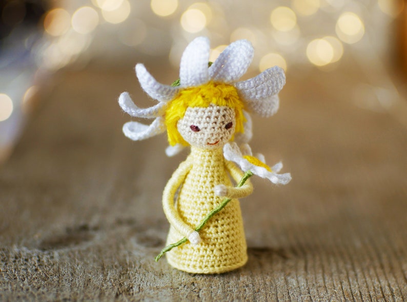 May include: A crocheted doll with yellow hair and a white daisy flower hat holding a green stem with a white daisy flower.