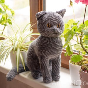 May include: A gray crocheted cat figurine with amber-colored eyes sits on a windowsill. The handmade cat has a textured surface and is positioned near potted plants. The cat's tail is extended, and the ears are perked up. The overall theme is home decor.