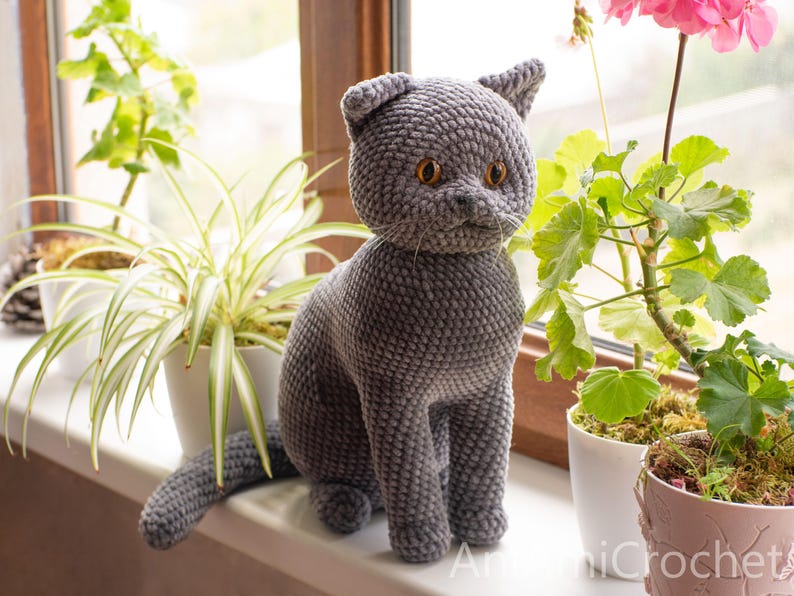 May include: A gray crocheted cat figurine with amber-colored eyes sits on a windowsill. The handmade cat has a textured surface and is positioned near potted plants. The cat's tail is extended, and the ears are perked up. The overall theme is home decor.