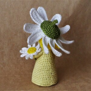 May include: A crocheted doll wearing a white daisy flower hat and holding a white daisy flower. The doll is wearing a yellow crocheted dress.
