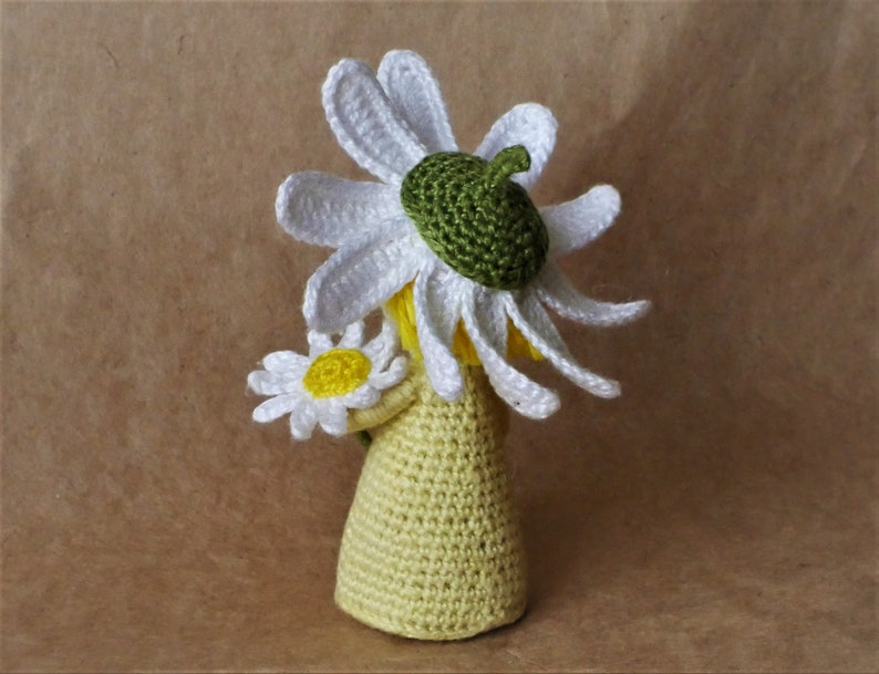 May include: A crocheted doll wearing a white daisy flower hat and holding a white daisy flower. The doll is wearing a yellow crocheted dress.