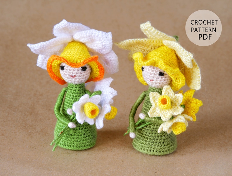 May include: Two crocheted daffodil dolls with green dresses and yellow and white flowers. The dolls have white and yellow crocheted hats. The dolls are on a brown surface. CROCHET PATTERN PDF.