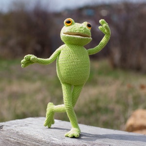 May include: A green crocheted frog toy with orange eyes, standing on a wooden surface with its right leg raised and its left arm outstretched.