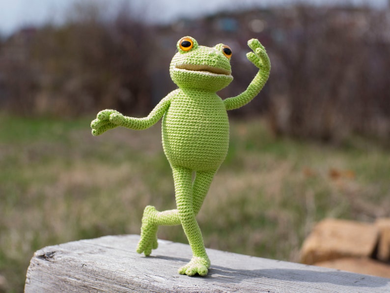 May include: A green crocheted frog toy with orange eyes, standing on a wooden surface with its right leg raised and its left arm outstretched.