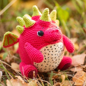 Op de afbeelding: Een gehaakt draken knuffel met roze en groene accenten. De draak heeft een wit en zwart gevlekte buik en een groene stekelige rug.