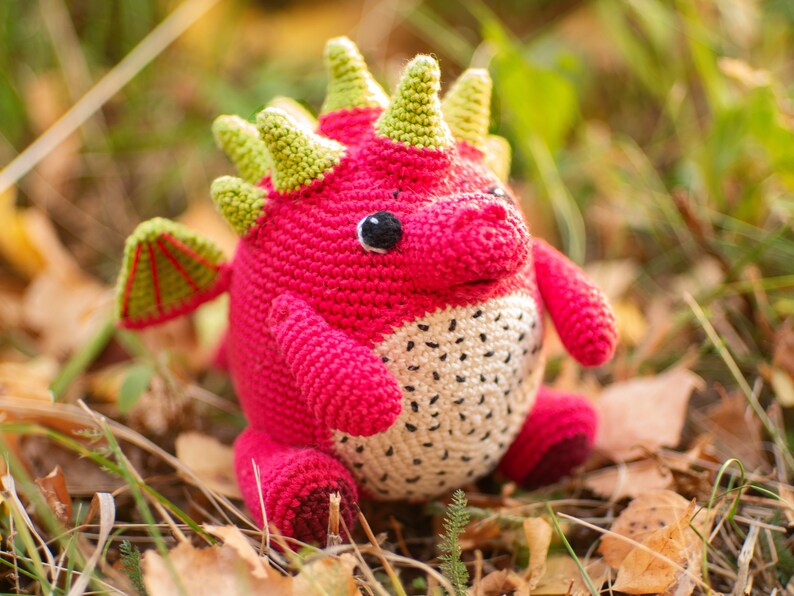 Op de afbeelding: Een gehaakt draken knuffel met roze en groene accenten. De draak heeft een wit en zwart gevlekte buik en een groene stekelige rug.