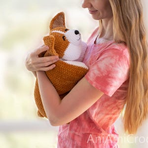 Peut inclure: Une peluche corgi crochet&eacute;e avec un corps brun dor&eacute; et un poitrail, un museau et des marques faciales blancs. Le jouet a des oreilles dress&eacute;es, un petit nez noir et des yeux ferm&eacute;s. Le texte AniAmiCrochet est visible dans le coin inf&eacute;rieur.