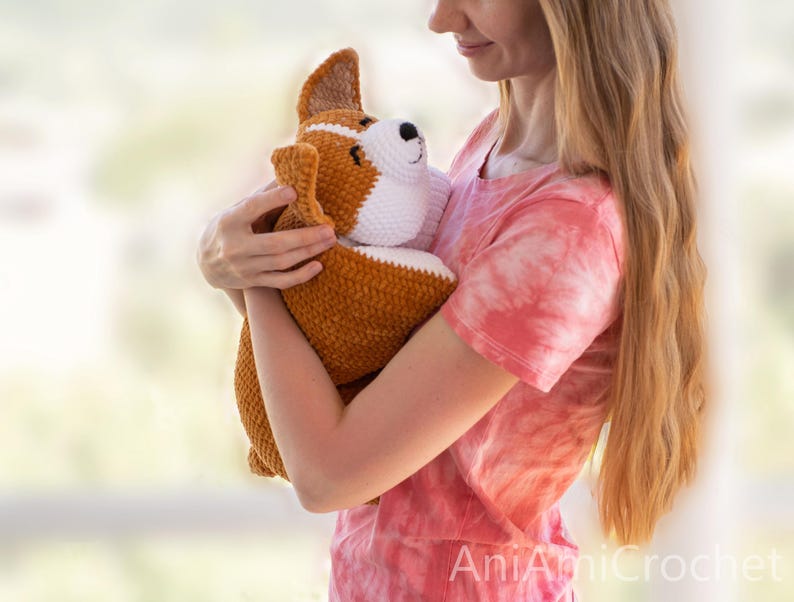 Peut inclure: Une peluche corgi crochet&eacute;e avec un corps brun dor&eacute; et un poitrail, un museau et des marques faciales blancs. Le jouet a des oreilles dress&eacute;es, un petit nez noir et des yeux ferm&eacute;s. Le texte AniAmiCrochet est visible dans le coin inf&eacute;rieur.