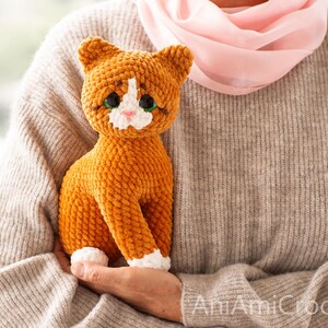 May include: A handmade orange and white crocheted cat plush toy with large dark green eyes, a small pink nose, and white patches on the face and paws. The cat has a textured, soft yarn appearance and is sitting in an upright position.