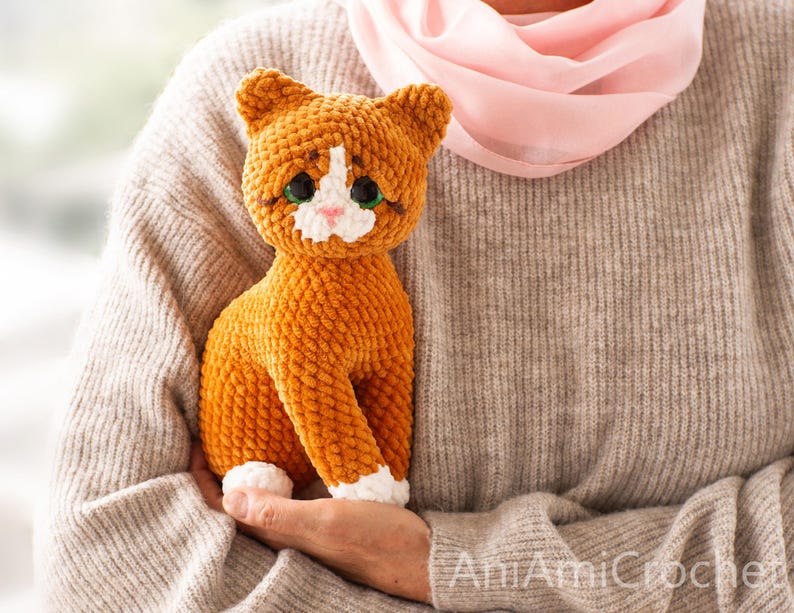 May include: A handmade orange and white crocheted cat plush toy with large dark green eyes, a small pink nose, and white patches on the face and paws. The cat has a textured, soft yarn appearance and is sitting in an upright position.
