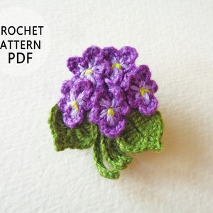 May include: Crochet pattern PDF for a purple and green crocheted flower bouquet. The flowers have yellow centers and are arranged in a cluster with green leaves.