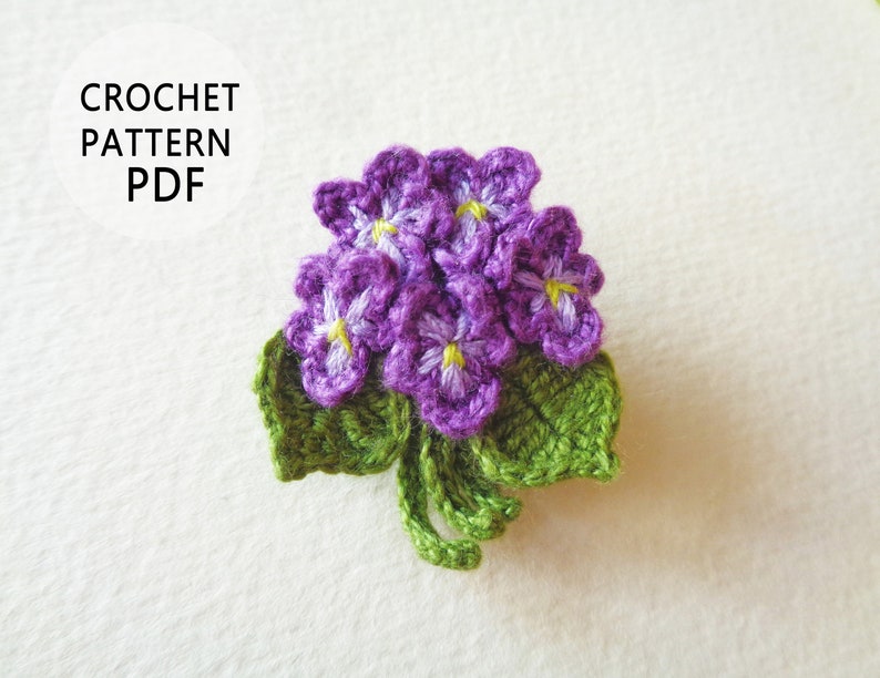 May include: Crochet pattern PDF for a purple and green crocheted flower bouquet. The flowers have yellow centers and are arranged in a cluster with green leaves.