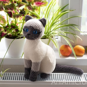May include: A crocheted Siamese cat plush toy featuring a cream-colored body with dark gray ears, face, paws, and tail. The cat has bright blue plastic eyes, stitched whiskers, and a small dark nose. The text AniAmicrochet is visible at the bottom.