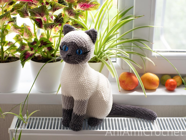 May include: A crocheted Siamese cat plush toy featuring a cream-colored body with dark gray ears, face, paws, and tail. The cat has bright blue plastic eyes, stitched whiskers, and a small dark nose. The text AniAmicrochet is visible at the bottom.
