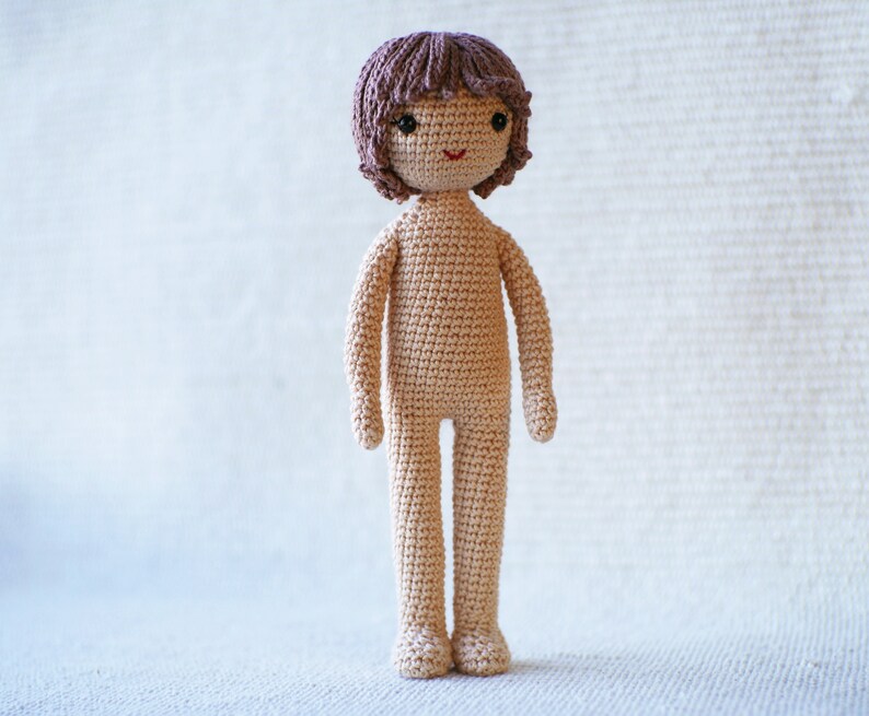 May include: A crocheted doll with brown hair, a beige body, and a smiling face.