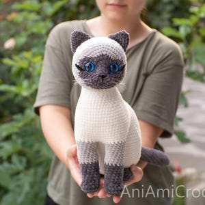 May include: A crocheted Siamese cat plush toy with a cream-colored body, dark gray ears, face mask, paws, and tail. The cat features bright blue plastic eyes, stitched whiskers, and the text AniAmiCrochet printed in the bottom right corner.