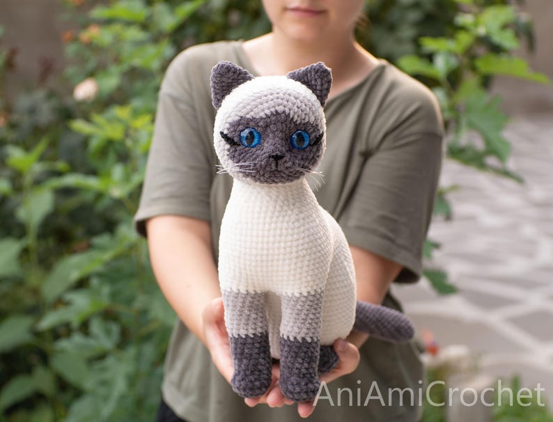 May include: A crocheted Siamese cat plush toy with a cream-colored body, dark gray ears, face mask, paws, and tail. The cat features bright blue plastic eyes, stitched whiskers, and the text AniAmiCrochet printed in the bottom right corner.